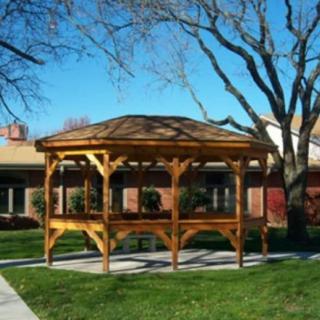 A wooden gazebo on a grassy lawn with trees and a building in the background.