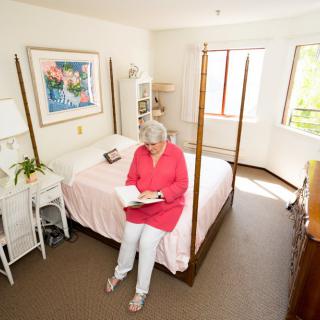 A woman sits on a bed in a brightly lit bedroom reading a book.