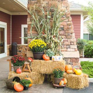 A festive autumn display featuring pumpkins, hay bales, flowers, and corn stalks in front of a brick wall.