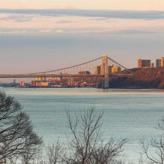 A scenic view of a bridge spanning across a river with a city skyline in the background during sunset.