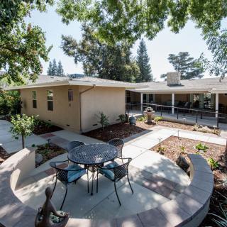 A modern patio area with a table and chairs surrounded by a landscaped garden and trees, adjacent to a house.