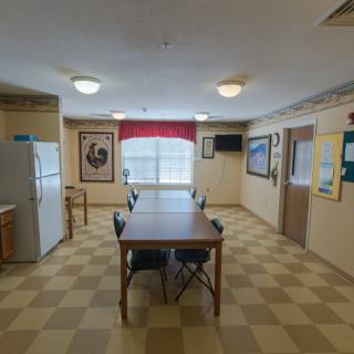 A communal kitchen space with a long table and chairs, featuring a refrigerator and artwork on the walls.