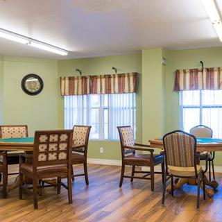 A well-lit room with tables and chairs arranged neatly, featuring windows with curtains and a wall clock.