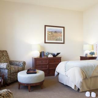 A cozy bedroom with an armchair, ottoman, dresser, and a neatly made bed.