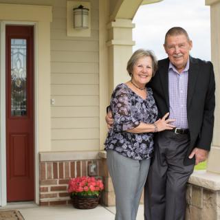 An elderly couple smiling and standing together in front of their home.