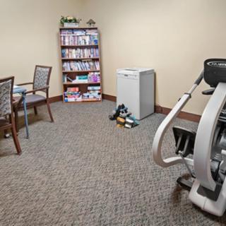 A small office space with an exercise machine, bookshelf, chairs around a table, and a mini fridge.