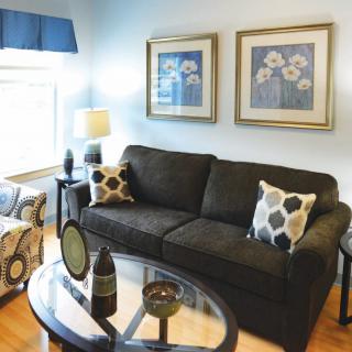A cozy living room with a dark sofa, patterned armchair, and glass coffee table, accented by framed artwork and decorative pillows.