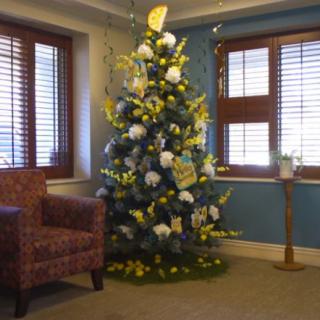 A decorated Christmas tree with blue and yellow ornaments stands in a cozy room next to a comfortable chair.