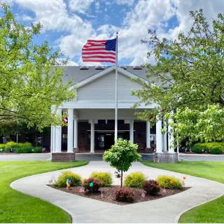 A building entrance with an American flag flying above, surrounded by well-maintained landscaping under a partly cloudy sky.