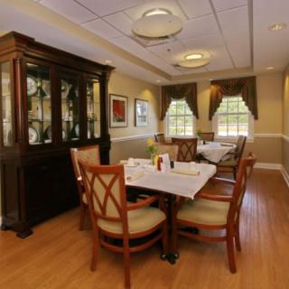 A cozy dining room with a neatly set table and chairs, accompanied by a wooden cabinet and sunlit windows.