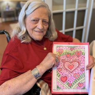 An elderly woman smiles while holding her colorful artwork featuring a heart design.