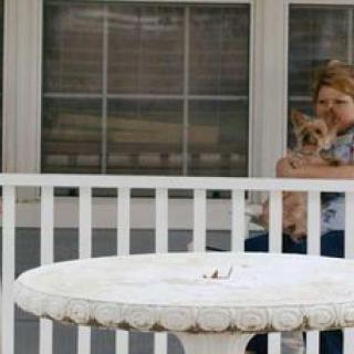 A woman sitting on a porch holding a dog, with a white table in the foreground.