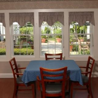 A dining area with a blue tablecloth and wooden chairs, set beside large windows that overlook a garden.