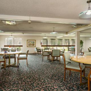 A spacious dining room with multiple tables and chairs, featuring patterned carpet and ceiling lights.