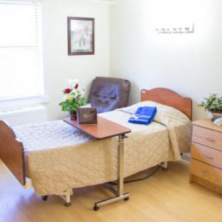 A neatly organized healthcare facility room featuring a single bed, a movable table, and comfortable seating.