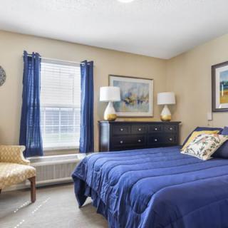 A cozy bedroom with a blue bedspread, decorated with artworks and featuring a wooden dresser and chair.