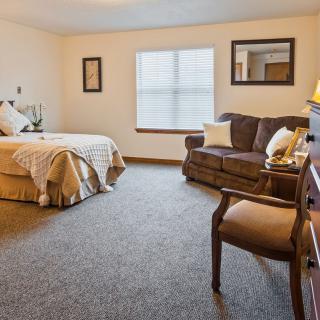 A cozy bedroom featuring a bed, sofa, and wooden chair with natural light coming through the window.