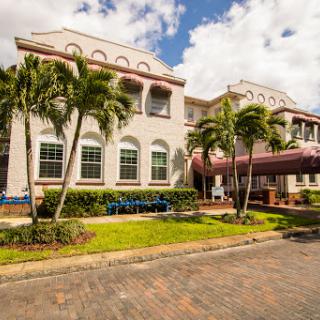 A beautiful architectural building surrounded by lush palm trees and landscaped gardens under a cloudy sky.