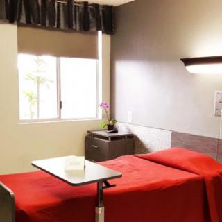 A tidy hospital room featuring a single bed with a red bedspread, a nightstand, and a window with curtains.