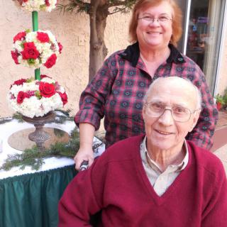 An elderly couple smiling outdoors with a floral arrangement in the background.