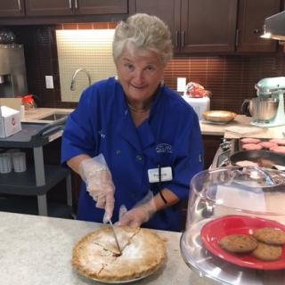 A woman in a blue shirt slicing a pie in the kitchen.