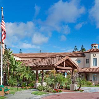 A Mediterranean-style building with an American flag in front, surrounded by lush landscaping under a clear blue sky.