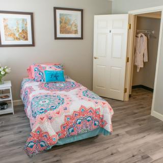 A cozy bedroom with a single bed featuring colorful bedding and decorative cushions, complemented by artwork on the walls and a wooden floor.