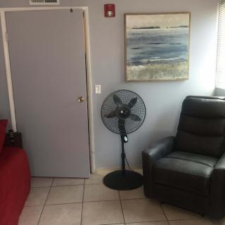 A cozy living space featuring a black armchair, a standing fan, and a seascape painting on the wall.