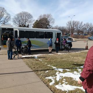 A group of people waiting at a bus stop on a chilly day, with some snow still visible.
