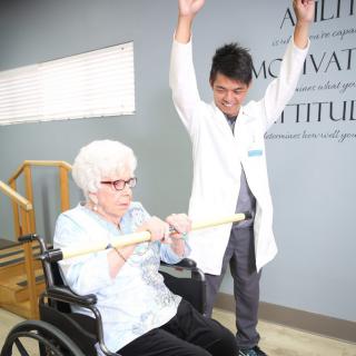 A healthcare professional assists an elderly woman in physical therapy exercises.