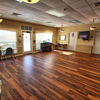 A spacious empty room with wooden flooring, featuring several chairs along the walls, a television mounted on the wall, and ample natural light from large windows.