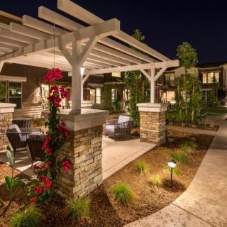 A beautifully lit outdoor seating area under a pergola, surrounded by lush landscaping and pathways at night.
