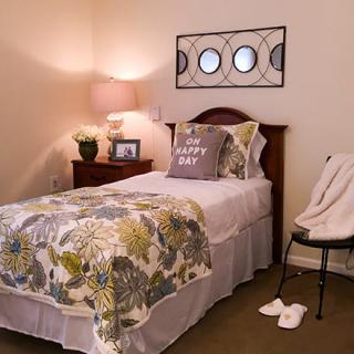 A cozy bedroom with a single bed featuring floral bedding, a nightstand with a lamp, and wall artwork.