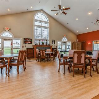 A spacious dining area with wooden floors and large windows, featuring multiple tables and chairs under a high ceiling.