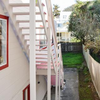 A view of a building with an external staircase leading to the upper level, adjacent to a narrow backyard with grass and a wooden fence.