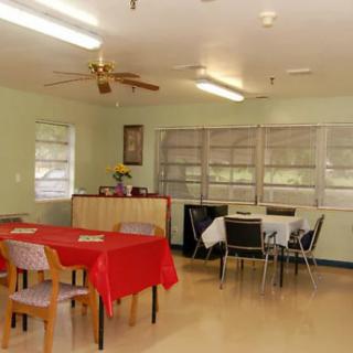 A simple dining room with tables covered in red and white tablecloths, surrounded by chairs and illuminated by ceiling lights.