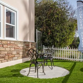 A small patio area with two metal chairs and a table on a round pebble surface, surrounded by a lush green lawn and a white picket fence.