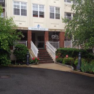 The image shows a building entrance with stairs leading up to the door, surrounded by landscaping with trees and bushes.