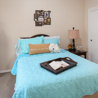 A cozy bedroom featuring a bed with blue bedding, a decorative throw pillow, and a tray holding an open book. A nightstand with a lamp adds to the room's warm ambiance.