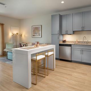 A modern kitchen with sleek gray cabinets, a compact island with bar stools, and light wood flooring.
