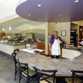 A chef stands behind a curved counter in a modern restaurant, with a buffet setup in the background.