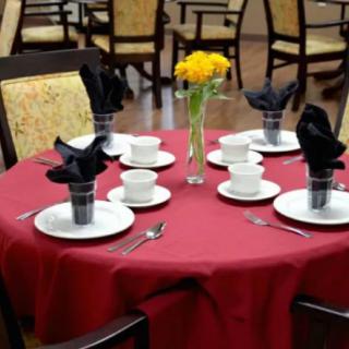 A neatly set dining table with red tablecloth, white dishes, black napkins, and a floral centerpiece.