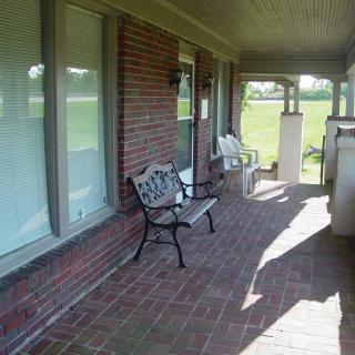 A shaded brick porch with benches and windows overlooking a lawn.