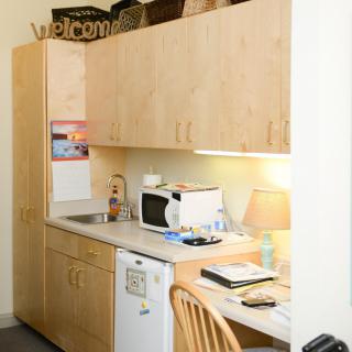 A compact kitchenette with wooden cabinets, a microwave, a small refrigerator, and a workspace with a desk lamp and notepad.