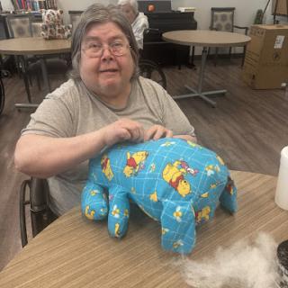 An elderly woman working on a colorful stuffed animal project at a crafting table.
