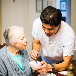 A caregiver assists an elderly woman by feeding her in a warm, home environment.