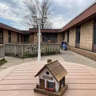 A serene courtyard featuring a miniature house in the foreground, surrounded by buildings and trees under a cloudy sky.