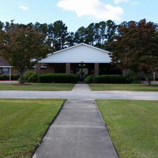 A building with a white facade is surrounded by trees and a well-maintained lawn, with a sidewalk leading to its entrance.