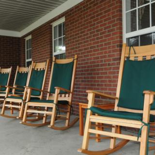 A row of wooden rocking chairs with green cushions on a porch of a brick building.