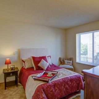 A cozy bedroom with red bedding and a small lamp on a nightstand by the bed, featuring a window providing natural light.